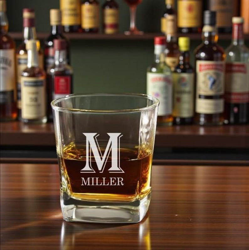 May include: A square-shaped, clear glass tumbler filled with amber liquid, likely whiskey. The glass is etched with a large white letter "M" above the name "MILLER". The background features a bar setting with bottles of liquor on shelves.
