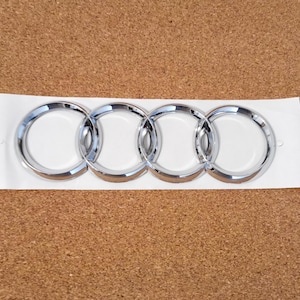 Peut inclure: Un emblème Audi chromé avec quatre anneaux entrelacés. L'emblème est sur un support blanc, posé sur une surface en liège marron. Les anneaux ont une finition brillante et réfléchissante.