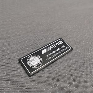 May include: A black and white AMG emblem with the text "AMG Mercedes-AMG AFFALTERBACH-GERMANY". The emblem has a rectangular shape with rounded corners. The emblem also features a circular logo with a tree.