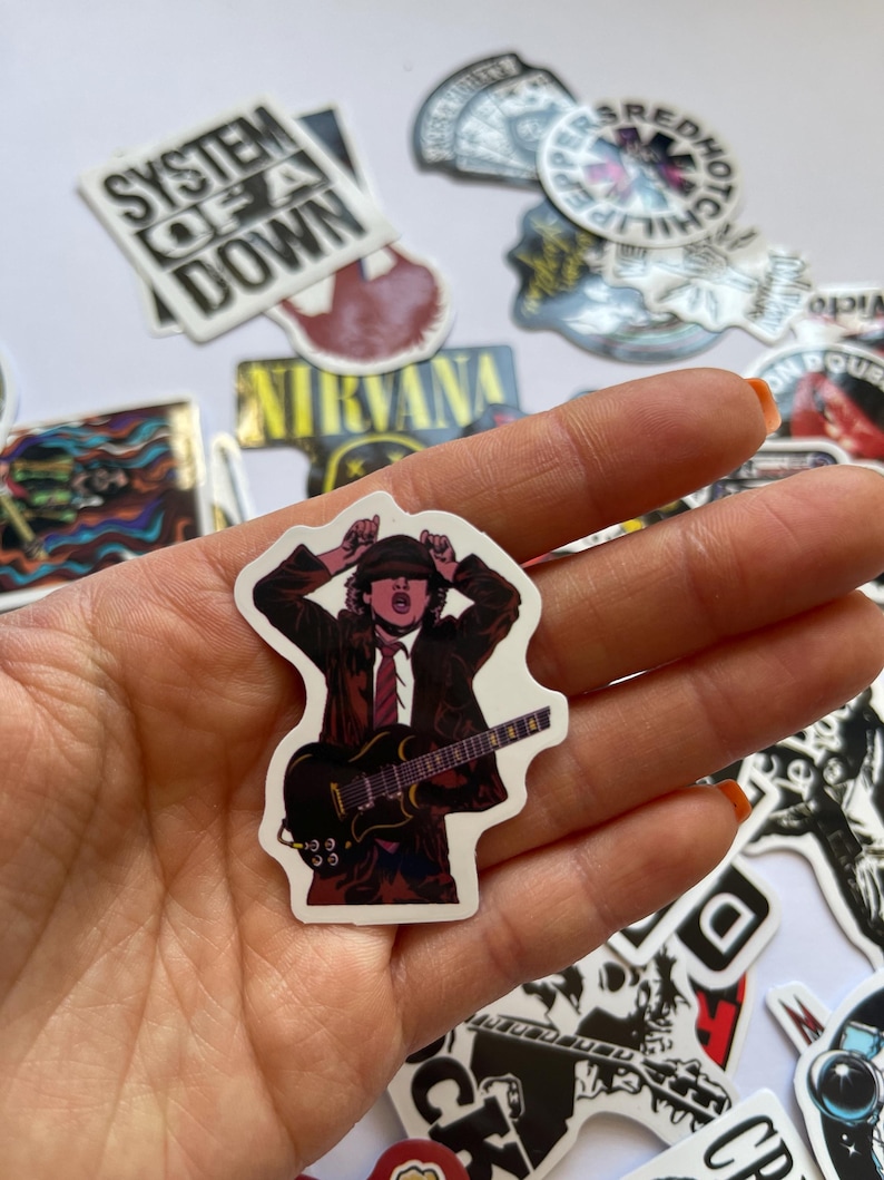 May include: A sticker featuring a cartoon illustration of a rock musician playing a guitar. The musician is wearing a black leather jacket, a red tie, and a black hat. The guitar is black with white accents. The sticker is on a white background.