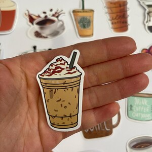 May include: A sticker of a iced coffee drink with whipped cream and a brown straw. The coffee is in a clear cup with a brown lid. The sticker is on a hand.