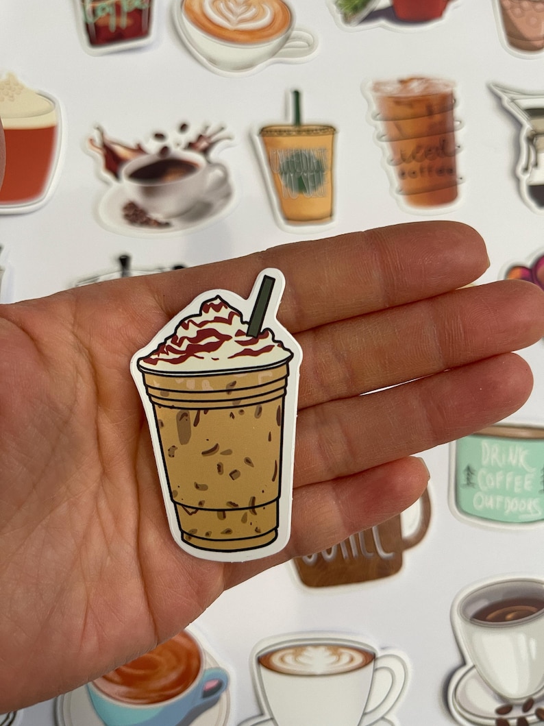 May include: A sticker of a iced coffee drink with whipped cream and a brown straw. The coffee is in a clear cup with a brown lid. The sticker is on a hand.