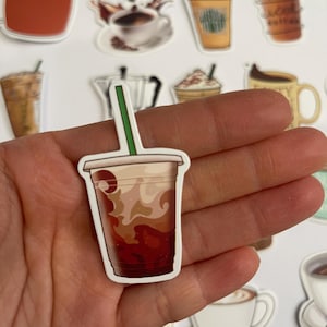 May include: A close-up of a sticker featuring a brown and white iced coffee drink in a clear plastic cup with a brown lid and a straw. The sticker is held in a hand.