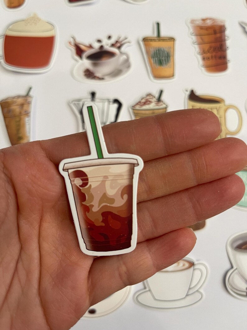 May include: A close-up of a sticker featuring a brown and white iced coffee drink in a clear plastic cup with a brown lid and a straw. The sticker is held in a hand.