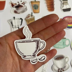 May include: A white sticker with a hand-drawn illustration of a coffee cup with steam rising from it and two coffee beans. The cup is brown and has a handle. The sticker is on a white background.
