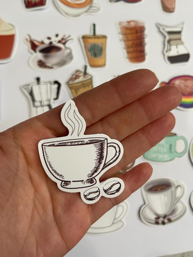 May include: A white sticker with a hand-drawn illustration of a coffee cup with steam rising from it and two coffee beans. The cup is brown and has a handle. The sticker is on a white background.