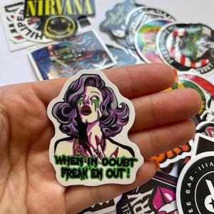 May include: A white sticker with a cartoon image of a woman with purple hair and green makeup. The text "WHEN IN DOUBT FREAK 'EM OUT!" is written in green letters on the sticker.