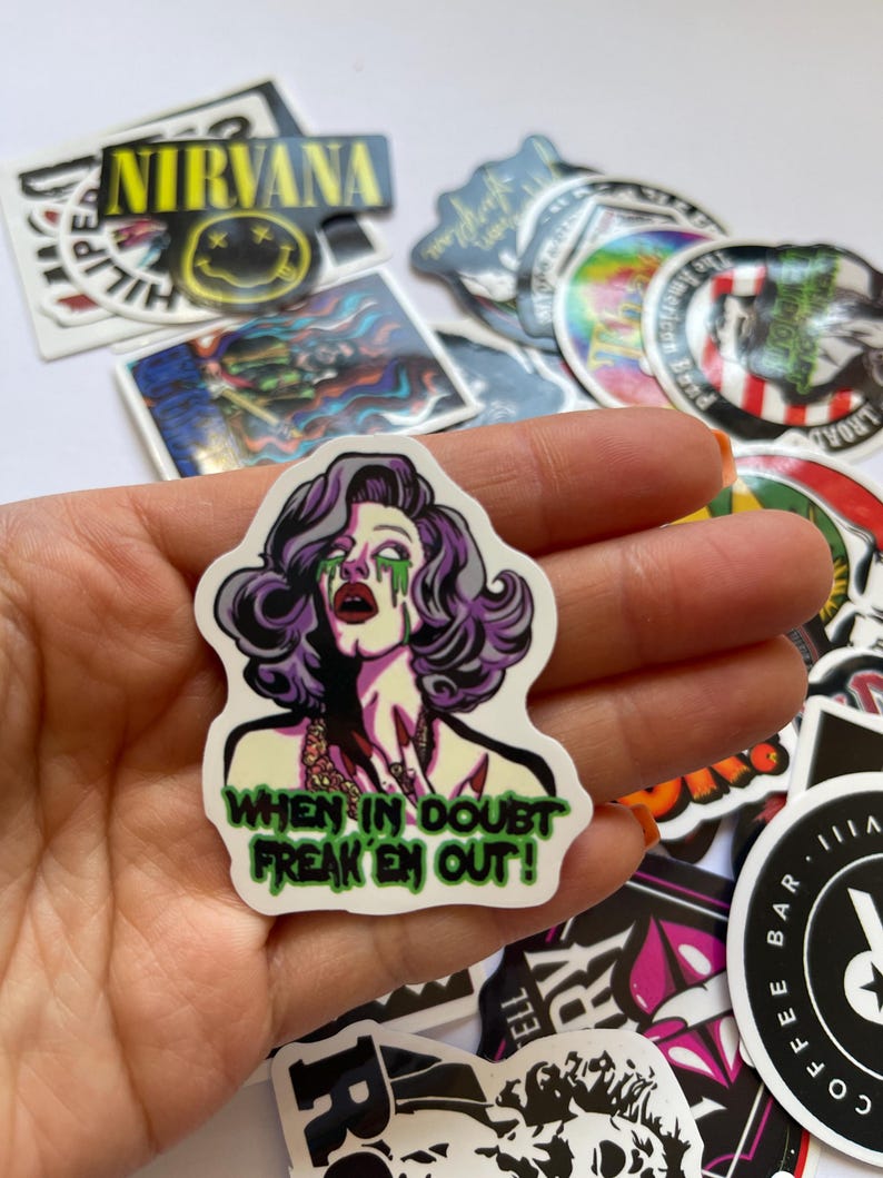 May include: A white sticker with a cartoon image of a woman with purple hair and green makeup. The text "WHEN IN DOUBT FREAK 'EM OUT!" is written in green letters on the sticker.