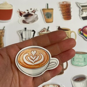 May include: A sticker of a latte with latte art in a white mug. The sticker is on a hand.