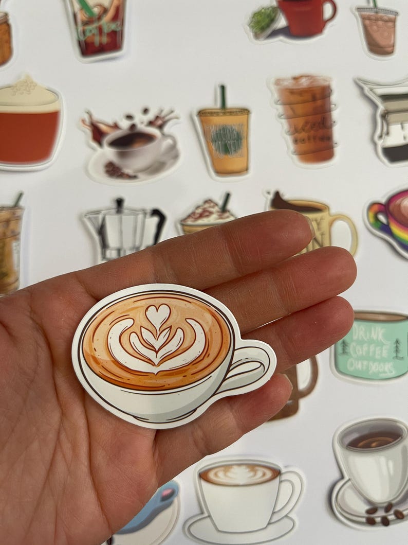 May include: A sticker of a latte with latte art in a white mug. The sticker is on a hand.