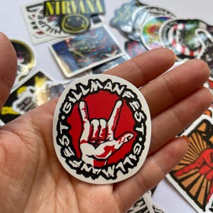 May include: A white sticker with a black circle around a red hand making the rock and roll hand gesture. The text "Gillmanfest" is written around the circle in black.