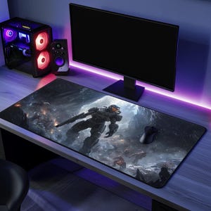 May include: A large rectangular gaming mousepad featuring a detailed illustration of a futuristic soldier in a battle scene. A black computer mouse sits on the right side. The scene includes a computer monitor, speaker, and a desktop computer.