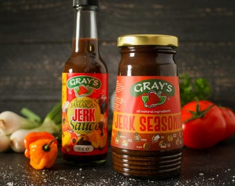 May include: Two bottles of Gray's Authentic Jamaican Jerk Sauce and Jerk Seasoning. The bottles are brown glass with red and white labels. The sauce bottle has a red pepper and the seasoning bottle has a red bell pepper on the label. The bottles are sitting on a dark wooden surface.