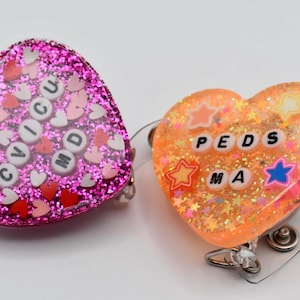 May include: Two heart-shaped resin badge holders with glitter and colorful accents. One is pink with white letters spelling "CIVICU MD" and the other is orange with white letters spelling "PEDS MA".