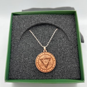 May include: A wooden pendant necklace with a silver chain, displayed in a green box. The pendant features a detailed star and eye design. The necklace is set against a black foam interior.