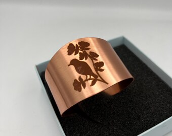 Può includere: Un bracciale a polsino in rame con un motivo a uccelli e fiori. L'ampio bracciale curvo è esposto in una scatola azzurra con interno nero. L'uccello è appollaiato su un ramo con foglie e fiori.
