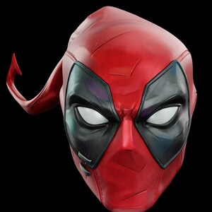 May include: A red and black Deadpool mask with white eyes and a distinctive headpiece. The mask has a textured surface and is set against a black background. The mask is a replica of the character's mask.