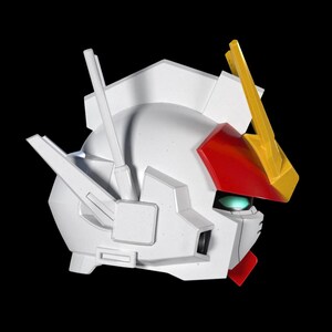 May include: A white robot head with red, yellow, and green accents. The head has a detailed design with angular features and a prominent crest. The robot head is set against a black background.