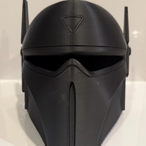May include: A black plastic helmet with a triangular design on the top and a visor. The helmet has a textured surface and appears to be 3D printed.