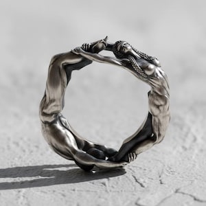 May include: A silver ring featuring two intertwined figures in a continuous loop. The ring's design showcases detailed human forms, with a matte finish. The figures appear to be embracing, creating a unique and artistic piece of jewelry.