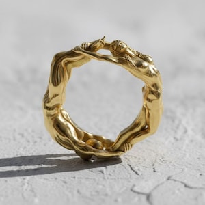 Op de afbeelding: Een goudkleurige ring met een uniek ontwerp. De ring heeft een doorlopende, verweven vorm, met twee figuren die elkaar bovenaan omarmen. De ring staat tegen een witte achtergrond, wat de details benadrukt.