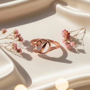 May include: A rose gold ring with the letter "D" in a cursive font. The ring is displayed on a white satin surface, with small pink flowers scattered around it. The ring is a delicate piece of jewelry.