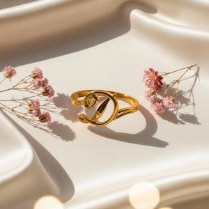 Op de afbeelding: Een goudkleurige ring met de letter 'D' in sierlijke letters. De ring ligt op een witte satijnen achtergrond, met kleine roze bloemen. Het ontwerp is elegant en modern, met een glanzende afwerking.