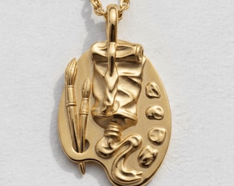 Può includere: Una collana con ciondolo color oro caratterizzata da un pendente ovale a forma di tavolozza da artista. Il ciondolo presenta un rilievo raffigurante un tubetto di colore, due pennelli e quattro piccole gocce di vernice, appeso a una catena.