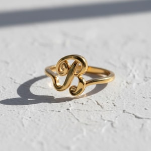 May include: A gold ring with the cursive letter "B" as its main design. The ring sits on a white, textured surface, with shadows adding depth. The ring is a piece of jewellery.