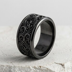 May include: A black ring with an intricate floral pattern. The ring is made of a dark metal and has a wide band. The design features swirling leaves and vines, creating a detailed and elegant look. The ring is a piece of jewelry.