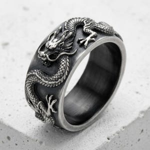 May include: A wide silver-toned metal ring featuring a detailed, raised relief of a dragon wrapping around the entire band. The surface has an oxidized, brushed finish that highlights the intricate scales, claws, and facial features of the dragon.