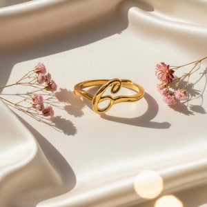 May include: A gold ring featuring the cursive letter 'C'. The ring is set against a white satin backdrop, with pink flowers. The ring is a piece of jewellery.