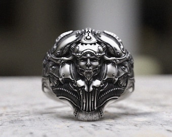 May include: A detailed silver ring with an intricate design. The ring features a central face with a beard, surrounded by ornate, mechanical-looking elements. The ring's design is symmetrical and has a futuristic, sci-fi aesthetic.