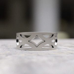 May include: A silver-colored ring with a geometric openwork design. The ring features a series of diamond and curved shapes cut out around the band. The ring is displayed on a light-colored surface.