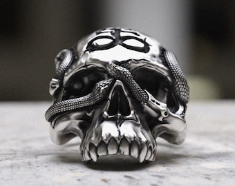 May include: A silver skull ring with a snake wrapped around its head. The snake's tail is curled around the skull's jaw. The skull has a detailed design with sharp teeth and a prominent brow.
