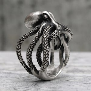 May include: A silver-toned ring shaped like an octopus with a textured head and multiple curved tentacles wrapping around the band. The metal features a polished finish with darkened recessed areas to highlight the circular suction cup details.