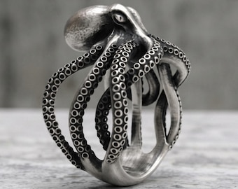 May include: A silver-toned ring shaped like an octopus with a textured head and multiple curved tentacles wrapping around the band. The metal features a polished finish with darkened recessed areas to highlight the circular suction cup details.