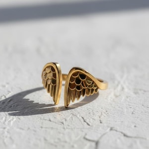 May include: A gold-colored ring with an open design featuring two stylized wing shapes. The ring is set against a white textured surface, with shadows adding depth. The jewelry piece is a fashion accessory.
