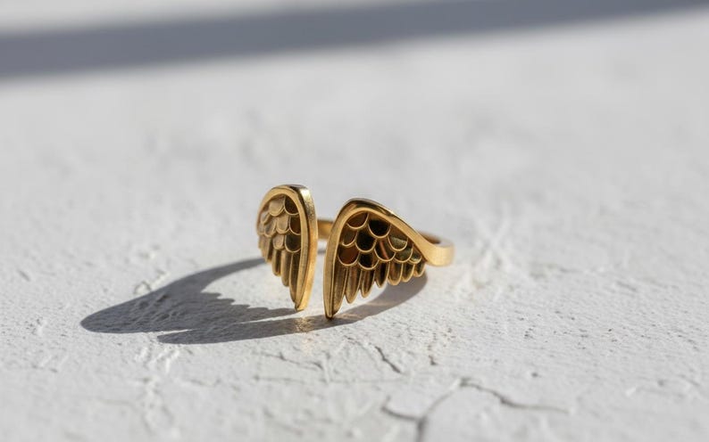 May include: A gold-colored ring with an open design featuring two stylized wing shapes. The ring is set against a white textured surface, with shadows adding depth. The jewelry piece is a fashion accessory.