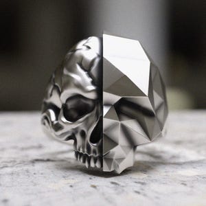 May include: A silver ring with a skull design on one side and a geometric design on the other side.