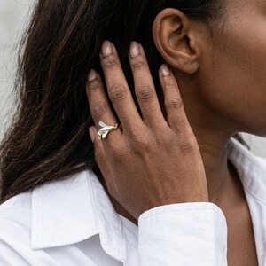 May include: A gold and silver ring with a bee design. The ring features a detailed bee with outstretched wings, set against a gold band. The ring is worn on a finger, showcasing the jewelry's craftsmanship and design.