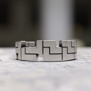 May include: A silver bracelet with a geometric pattern. The bracelet is made of interlocking pieces that form a repeating design.