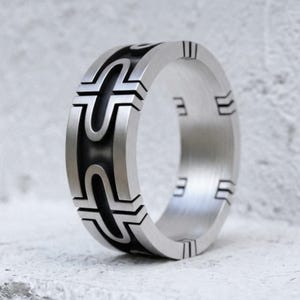 May include: A wide silver-toned metal ring featuring a brushed finish and a central black recessed band. The exterior surface displays a repeating geometric pattern of interlocking U-shaped lines and vertical notches that wrap around the band.