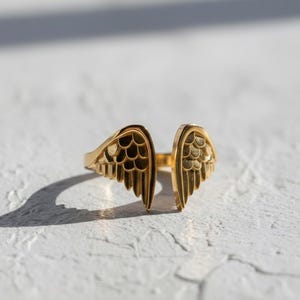 May include: A gold-colored ring with an open design, featuring wing-shaped details on each side. The ring is set against a white textured surface, with shadows adding depth. The wings have intricate feather detailing.