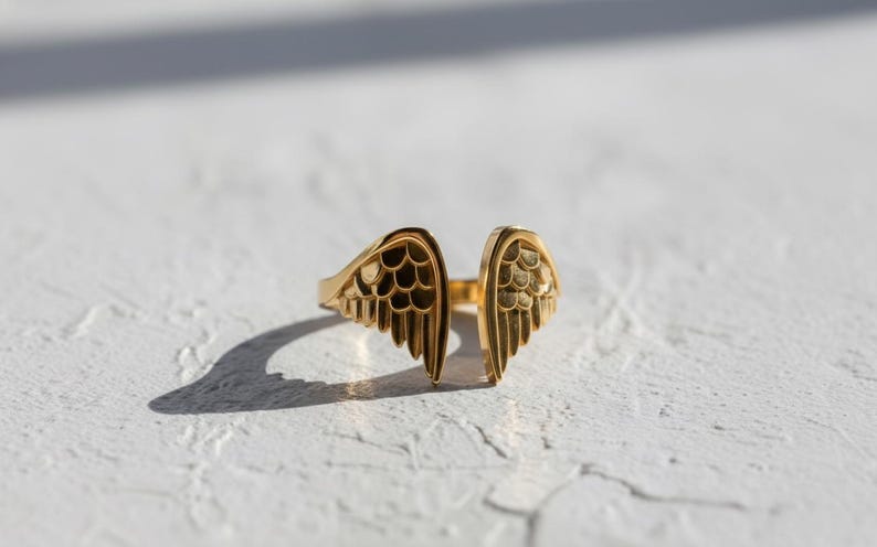 May include: A gold-colored ring with an open design, featuring wing-shaped details on each side. The ring is set against a white textured surface, with shadows adding depth. The wings have intricate feather detailing.