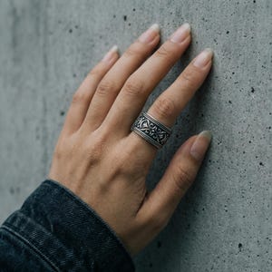 Könnte beinhalten: Ein silberfarbener Ring mit einem aufwendigen, sich wiederholenden Muster. Der Ring ist breit und sitzt auf einem Finger, wodurch das detaillierte Design zur Geltung kommt. Die Oberfläche des Rings reflektiert das Licht und hebt die geprägten Details hervor. Der Ring ist ein Schmuckstück.