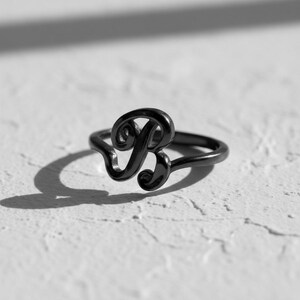 Può includere: Un anello in metallo nero con la lettera "B" in corsivo. L'anello è posto su una superficie bianca strutturata, con un'ombra proiettata a sinistra. È un gioiello.