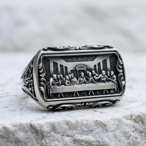Può includere: Un anello in metallo color argento con una faccia rettangolare caratterizzata da un rilievo dettagliato dell'Ultima Cena. La scena raffigura figure sedute a un lungo tavolo, incorniciate da motivi ornamentali a volute sui lati dell'anello.