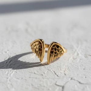 May include: A gold-colored ring with an open design, featuring two detailed wing shapes. The ring is set against a white textured surface, with shadows adding depth. The jewelry piece is a stylish accessory.