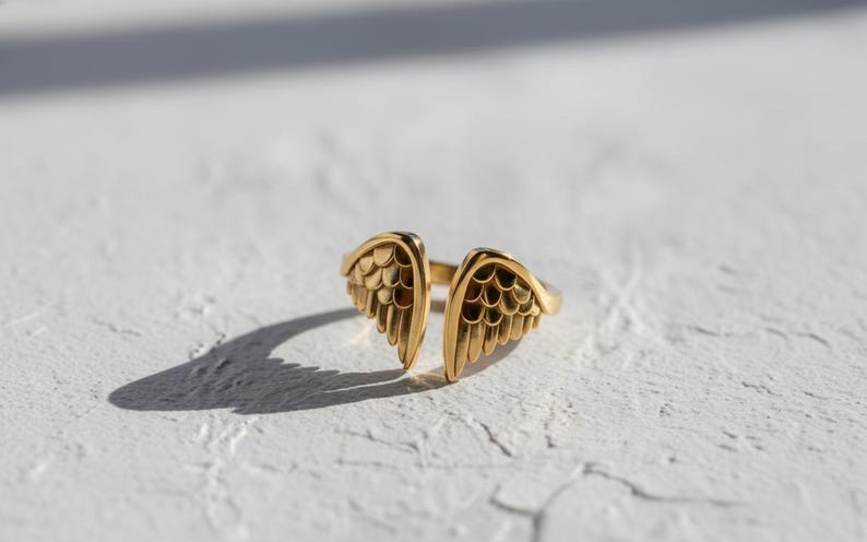 May include: A gold-colored ring with an open design, featuring two detailed wing shapes. The ring is set against a white textured surface, with shadows adding depth. The jewelry piece is a stylish accessory.